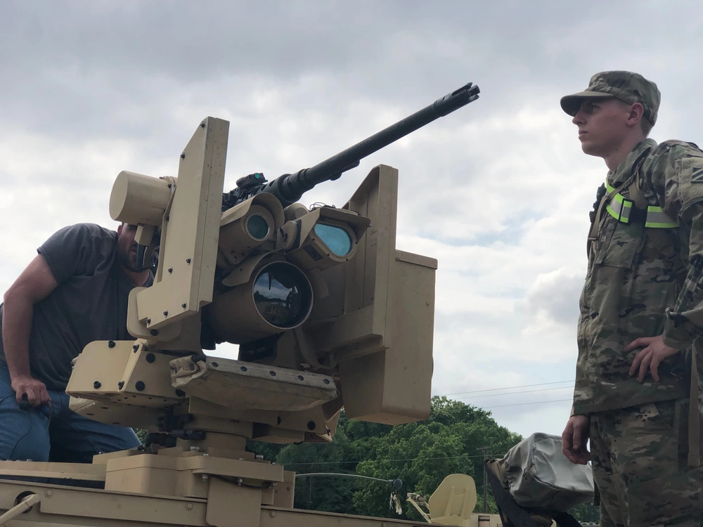 The image shows two individuals on top of a military vehicle examining a Remote Weapon Station (RWS) mounted with what appears to be a medium-caliber autocannon. One person is in US Army OCP (Operational Camouflage Pattern) uniform, while a civilian in casual clothing inspects the weapon system up close. The setting appears to be an outdoor demonstration or static display event based on the overcast sky and surrounding trees.