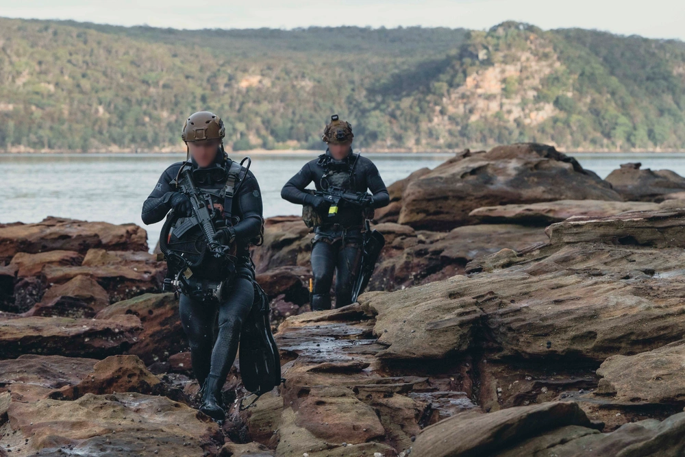 U.S. Naval Special Warfare (NSW) operators navigate over rocky terrain on the shoreline during joint over-the-beach training with Marine Raiders assigned to U.S. Marine Forces Special Operations Command as part of Exercise Talisman Sabre 25 in Sydney, Australia, July 15, 2025. Credit: Mass Communication Specialist 2nd Class David Rowe