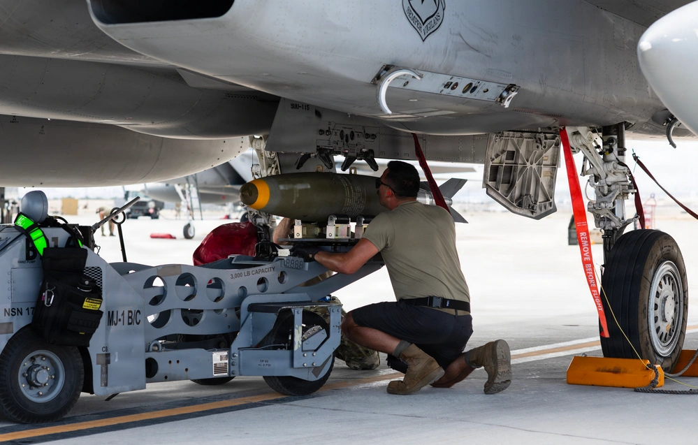 142nd Wing F-15EXs drop Live Bombs in Historic First A military aircraft maintainer is loading or securing a munition onto the underside of a military jet aircraft on a flight line. The technician is using an MJ-1B/C weapons loading cart, rated at 3,000 lb capacity, to position what appears to be a bomb or guided munition onto a hardpoint beneath the aircraft. Safety equipment including red 'Remove Before Flight' streamers and wheel chocks are visible.