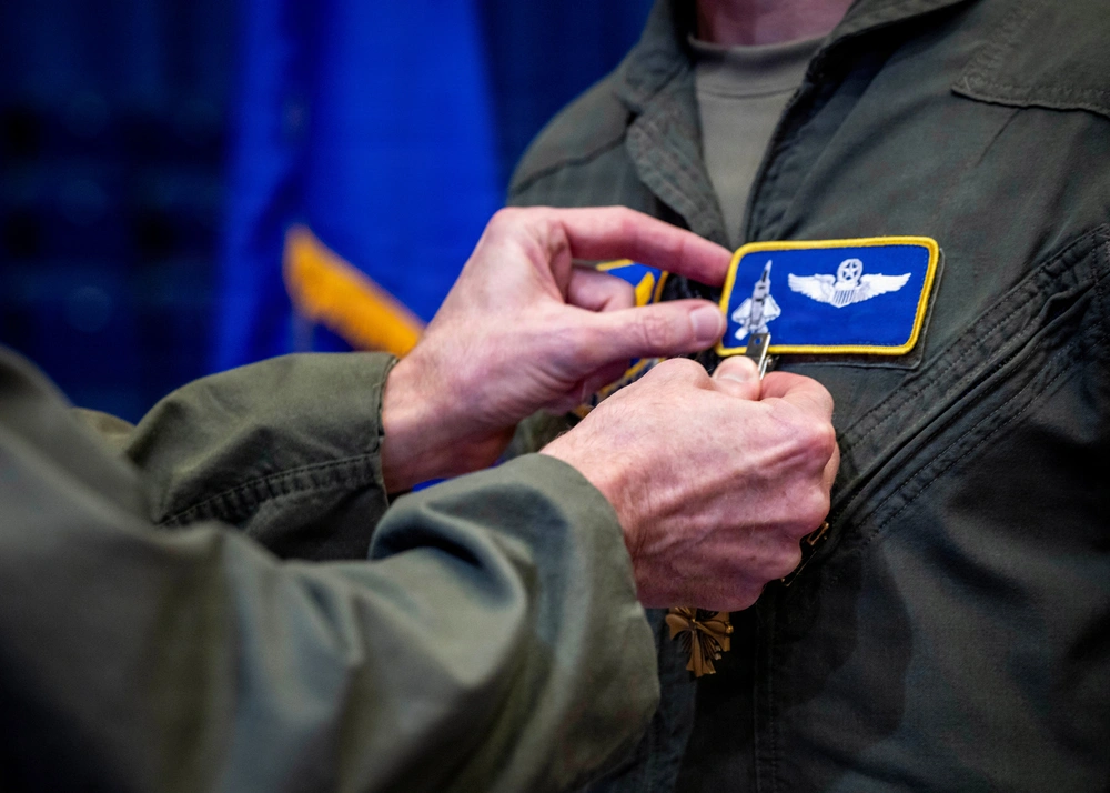 U.S. Air Force Pilot Receives Distinguished Flying Cross After Shielding Strike Package A close-up photograph showing a military pinning ceremony where one person is attaching a medal or badge to another person's flight suit. The recipient is wearing an olive green flight suit with a blue and gold patch depicting a military aircraft and aviator wings. This appears to be a formal military recognition or award ceremony.